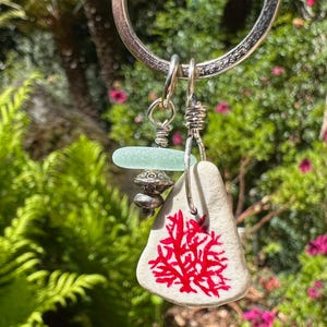May include: A silver keychain with a sea-themed charm. The charm features a white, triangular piece with a red coral design, a light blue glass bead, and silver beads. The background is a blurred view of green foliage and pink flowers.