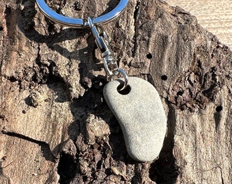 Könnte beinhalten: Ein glatter, grauer Stein-Schlüsselanhänger mit einem silbernen Metallring und einer Kette. Der Stein ist nierenförmig und hat ein durchgehendes Loch.