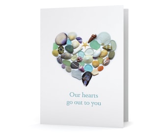 May include: A white greeting card featuring a heart shape made of seashells, sea glass, and a starfish. The text "Our hearts go out to you" is printed in teal below the heart. The card is folded.