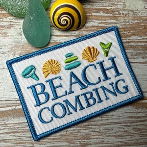 Puede incluir: Parche bordado con las palabras "BEACH COMBING" en azul, rodeado por un borde azul. El parche presenta imágenes bordadas de conchas marinas y otros objetos con temática de playa en tonos turquesa y dorado.