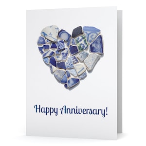 May include: A white greeting card with a heart made of blue and white ceramic shards. The words "Happy Anniversary!" are written in navy blue script below the heart. The card is folded.