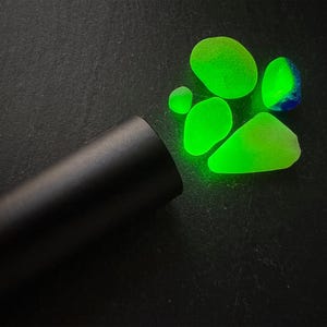 May include: A black flashlight illuminates a cluster of glowing green sea glass pieces on a dark surface. The sea glass varies in size and shape, with one piece showing blue hues. The scene suggests a treasure hunt or a display of unique finds.