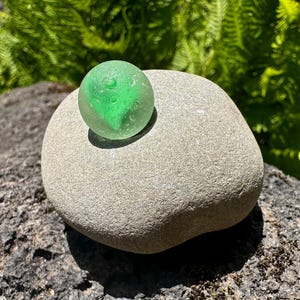 Op de afbeelding: Een gladde, bolvormige, doorschijnend groene glazen knikker met een matte, verweerde textuur die op een afgeronde, lichtgrijze, getextureerde riviersteen ligt. De knikker vertoont subtiele interne variaties en een matte afwerking.