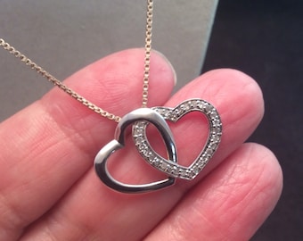 May include: A silver-coloured necklace featuring two interlocking heart-shaped pendants. One heart has a smooth, polished finish, whilst the other is encrusted with small, clear stones along its front surface. The pendant hangs from a delicate box chain.