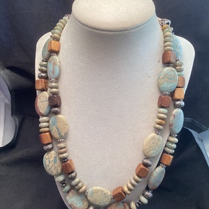 May include: A double-strand necklace with oval-shaped green and brown stones and wooden beads. The necklace is displayed on a white mannequin.