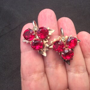 May include: A pair of vintage earrings featuring clusters of bright red, round gemstones. The earrings have a gold-toned metal leaf design with a small, clear accent stone. The earrings are shown against a dark background.
