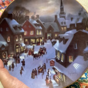May include: Decorative plate featuring a winter scene of a historic town square. The artwork depicts snow-covered buildings, horse-drawn carriages, and figures in period clothing. The plate has a gold rim and is held by a person.