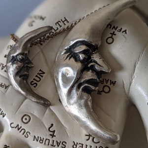 May include: A silver necklace with two crescent moon pendants, each with a face. The necklace is on a white surface with astrological symbols and text, including "JUPITER SATURN SUN", "MARS", "VENUS", and "HEALTH".