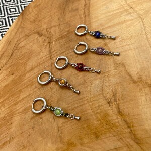 Peut inclure: Cinq boucles d'oreilles créoles argentées, chacune dotée d'une petite breloque pendante. Les breloques se composent d'une perle ronde centrale bleue, rose, rouge, jaune ou verte, entourée d'un cadre en fil tressé avec une pointe métallique.