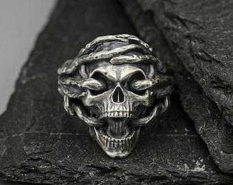 May include: A silver skull ring with a detailed design of a skull with a crown of thorns. The ring is on a black slate background.