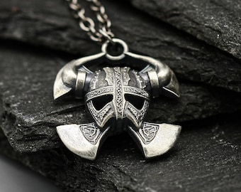 May include: Silver pendant necklace featuring a Viking helmet with two axes on either side. The pendant is on a black slate background.