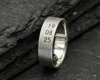 May include: A silver ring with a textured surface, engraved with the numbers "19 08 25". The ring is set against a dark, textured background, creating a contrast that highlights the ring's details. The ring is a simple band design.