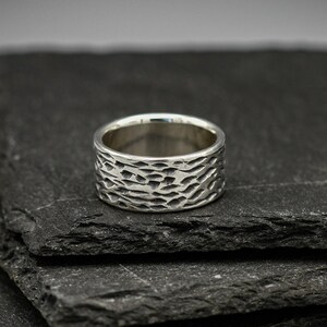 Puede incluir: Un anillo de plata con un acabado texturizado martillado. El anillo se muestra en una pila de baldosas de pizarra negra.