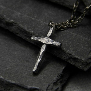 May include: A silver cross pendant necklace with a hammered texture, hanging on a black chain. The necklace is displayed on a stack of black slate tiles.