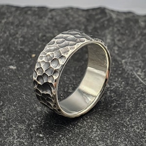 Könnte beinhalten: Ein silberner Ring mit gehämmerter Textur. Der Ring ist breit und hat eine glatte Innenfläche.