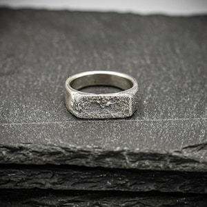 Op de afbeelding: Een zilveren ring met een rechthoekige voorkant en een getextureerd oppervlak. De ring staat tegen een donkere, gelaagde achtergrond, wat de metaalglans benadrukt. Het ontwerp van de ring is eenvoudig maar opvallend, geschikt voor dagelijks gebruik.