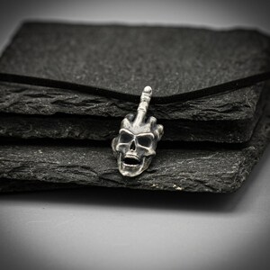 May include: A silver pendant necklace featuring a skull with a middle finger raised in the air. The pendant is on a black cord and is resting on a stack of black slate tiles.