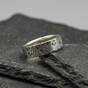 May include: A silver ring with a textured, patterned design. The ring is sitting on a stack of black slate tiles.