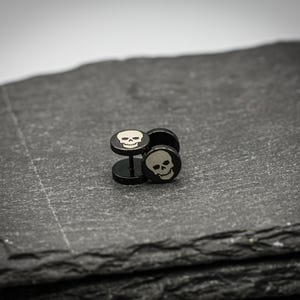 Puede incluir: Pendientes de calavera negros con diseño de calavera plateada. Los pendientes se muestran sobre una superficie gris oscura y texturizada. Los pendientes tienen un poste y una cara plana y redonda con una imagen de calavera.