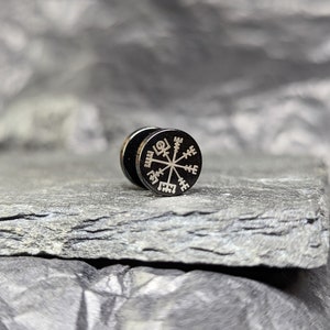 Puede incluir: Un pendiente de metal negro con un diseño de brújula plateado. La brújula presenta un círculo con ocho puntos y un símbolo en el centro.