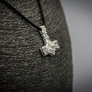 Puede incluir: Un collar con un colgante de plata con un diseño de martillo Mjolnir texturizado. El collar está en un cordón de cuero negro.