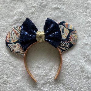 May include: A pair of blue and gold sequined Minnie Mouse ears with a seashell print on the ears. The headband is a light brown color.