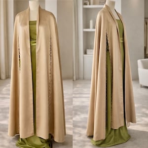 May include: A floor-length beige satin cape featuring a structured collar and a vertical row of small buttons along each side opening, worn over a sleeveless olive green satin gown with a fitted bodice and a long, flowing skirt silhouette.
