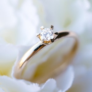May include: A gold engagement ring with a single, clear diamond set on a white flower. The ring's band is thin and polished, reflecting light. The diamond is the focal point, sparkling against the soft, blurred background.