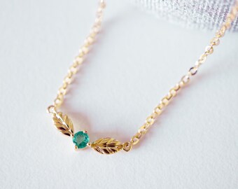 May include: A delicate gold necklace featuring a small, round, emerald-green gemstone. The pendant is flanked by two gold leaf designs. The necklace chain is a fine, linked style, and the background is a soft, neutral color.