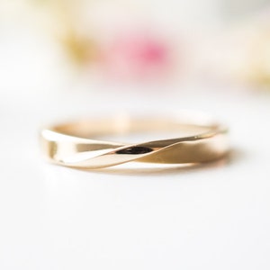 May include: A gold ring with a twisted design. The ring has a smooth, polished surface. The background is blurred, with soft pink and green hues. The ring's design is a continuous loop.