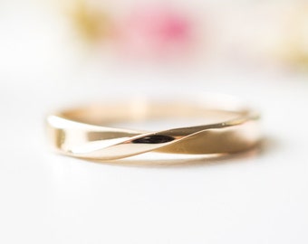 May include: A gold-colored ring with a twisted design. The ring is smooth and shiny, reflecting light. The ring is set against a soft, blurred background with hints of pink and green.