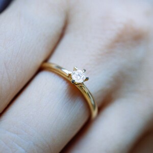 May include: A delicate gold ring with a single, sparkling diamond set in a six-prong setting. The ring is on a finger, showcasing its simple elegance and classic design. Ideal for engagement or everyday wear.