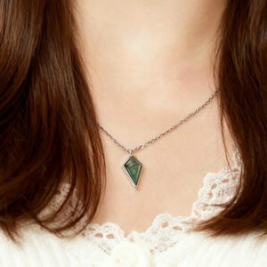 May include: A silver necklace featuring a diamond-shaped pendant with a green stone. The pendant is framed in silver and hangs from a delicate chain. The necklace is worn against a white lace top.