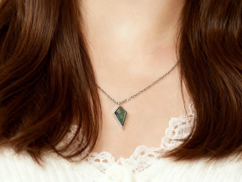 May include: A silver necklace featuring a diamond-shaped pendant with a green stone. The pendant is framed in silver and hangs from a delicate chain. The necklace is worn against a white lace top.