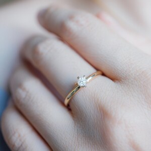 May include: A gold ring with a single, round, clear diamond set in a four-prong setting. The ring is on a finger, with the hand and arm in the background.