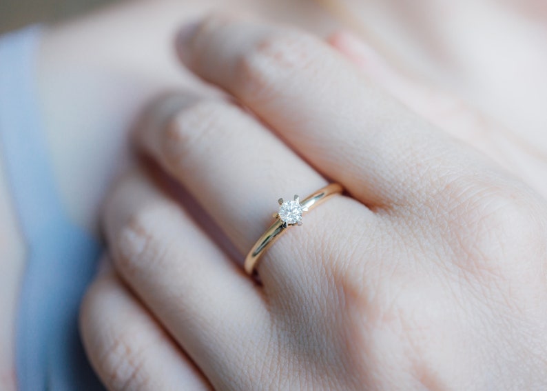May include: A gold ring with a single, round, clear diamond set in a four-prong setting. The ring is on a finger, with the hand and arm in the background.