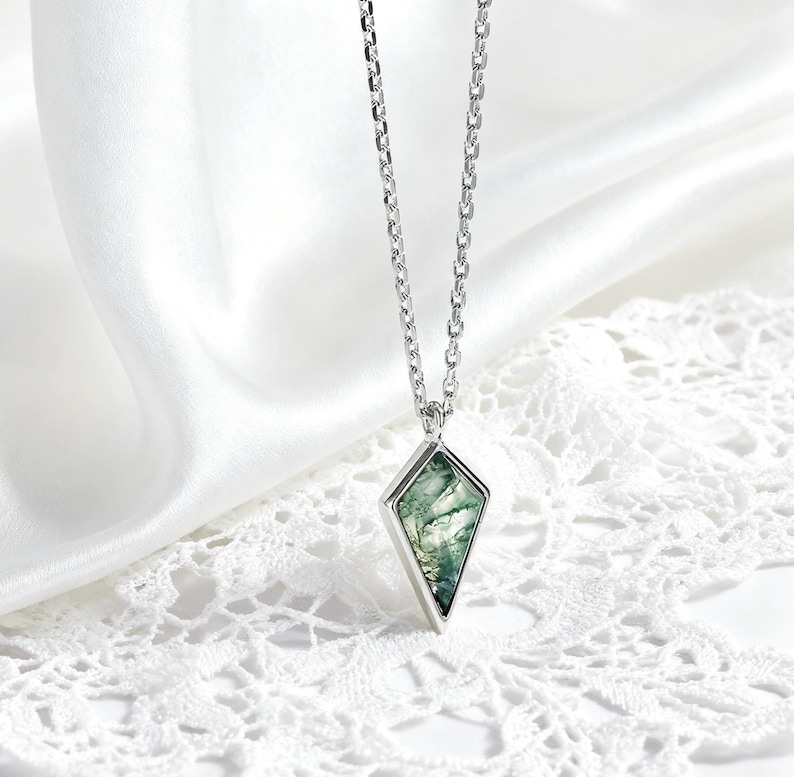 May include: A silver necklace with a diamond-shaped pendant. The pendant features a green and white stone set in a silver frame. The necklace hangs on a delicate silver chain, displayed on a white lace surface.