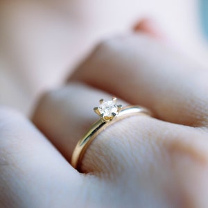 May include: A gold ring with a small, clear diamond set in a four-prong setting. The ring is simple and elegant, with a thin band. The ring is on a finger.