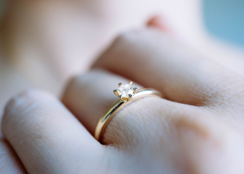 May include: A gold ring with a small, clear diamond set in a four-prong setting. The ring is simple and elegant, with a thin band. The ring is on a finger.