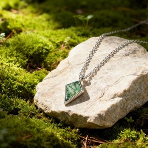 May include: A silver necklace with a geometric pendant featuring a green stone. The pendant is diamond-shaped with a silver border. The necklace rests on a light-colored rock surrounded by green moss.