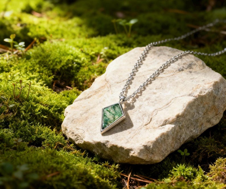 May include: A silver necklace with a geometric pendant featuring a green stone. The pendant is diamond-shaped with a silver border. The necklace rests on a light-colored rock surrounded by green moss.