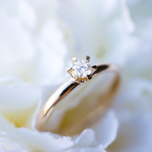 May include: A gold ring with a single, clear diamond set in a six-prong setting. The ring is set against a soft, white background, creating a contrast that highlights the ring's details. The ring is a symbol of commitment and love.