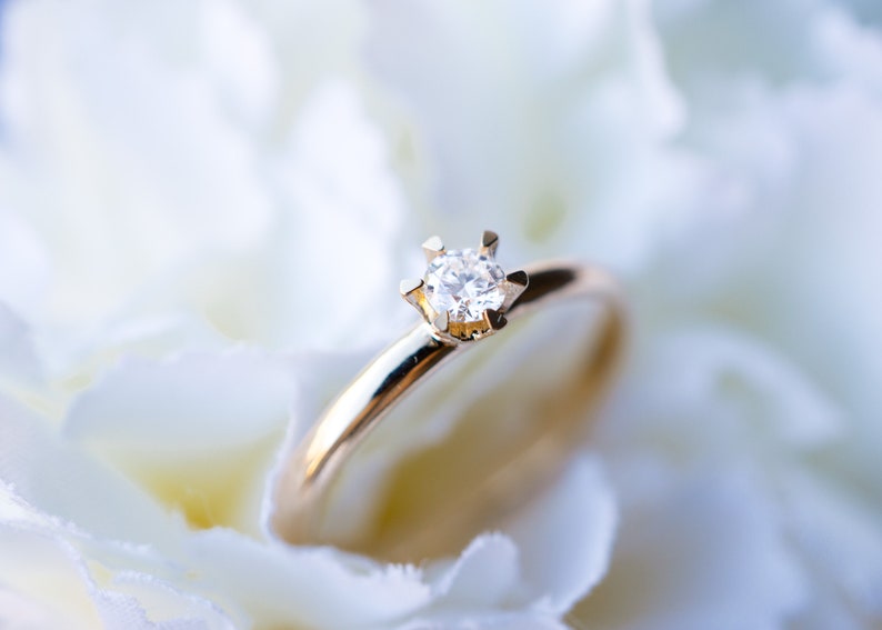 May include: A gold ring with a single, clear diamond set in a six-prong setting. The ring is set against a soft, white background, creating a contrast that highlights the ring's details. The ring is a symbol of commitment and love.