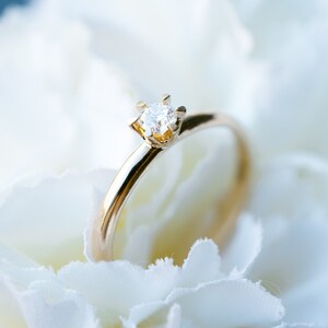 May include: A gold ring with a single, clear diamond set in a four-prong setting. The ring is displayed on a bed of white petals, creating a soft, romantic aesthetic. The ring's band is smooth and polished.