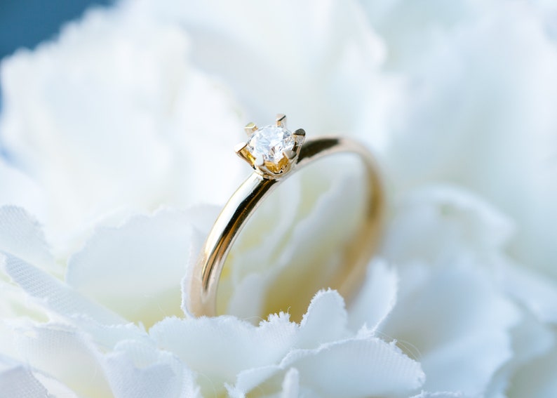 May include: A gold ring with a single, clear diamond set in a four-prong setting. The ring is displayed on a bed of white petals, creating a soft, romantic aesthetic. The ring's band is smooth and polished.