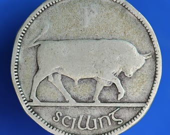 May include: A silver coin featuring a bull walking to the left. The coin has a serrated edge and the word "Scilling" is inscribed at the bottom. The number "1" and the letter "S" are at the top. The coin is on a blue background.