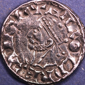 May include: A silver coin with a design of a stylized animal with a crown on its head. The coin has a circular shape and a raised rim. The coin is surrounded by a circular inscription.