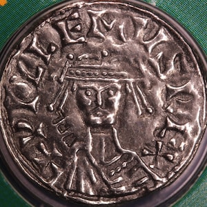 May include: A silver coin with a portrait of a crowned figure on one side. The coin has a raised rim and a smooth surface.