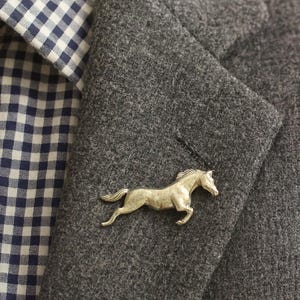 May include: A silver-toned metal brooch shaped like a galloping horse, pinned to the lapel of a dark grey wool-textured blazer worn over a navy blue and white gingham patterned collared shirt with a button-down collar and visible stitching.