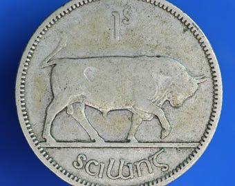 May include: A silver coin featuring a bull walking to the right. The coin has the text "1s" at the top and "Scilling" at the bottom. The coin has a textured edge and is set against a blue background.